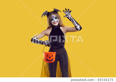 Happy child in spooky Halloween costume holding candy basket, waving hello and smiling 93316173