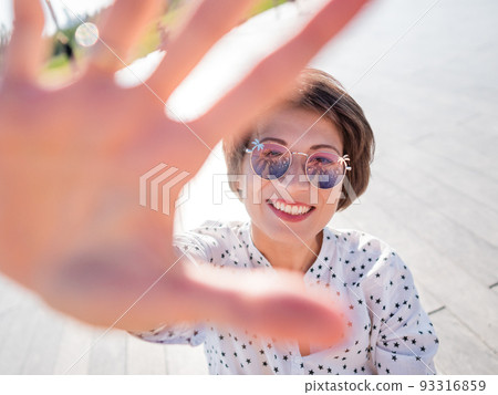 Wind ruffles short hair of freckled woman in colorful sunglasses. Smiling woman at open wooden scene of urban park. Summer vibes. Sincere emotions. 93316859