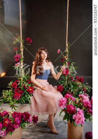 a beautiful young woman in a dress sits on a swing in peony flowers. solar glare 93318347