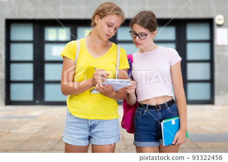 Portrait of female school students with notebooks speaking while standing on street in good mood near school. Friends concept Portrait of female school students with notebooks speaking while standing on street in good mood near school. Friends concept 93322456