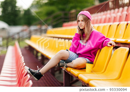 A teenage girl is sitting at the stadium at halftime and talking on the phone with friends 93330094