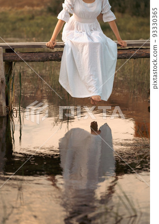 Barefoot woman in vintage white dress sitting on wooden rustic bridge over the little river, at sunset, faceless, vertical photo, selective focus. Barefoot woman in vintage white dress sitting on wooden rustic bridge over the little river, at sunset, faceless, vertical photo, selective focus. 93330365