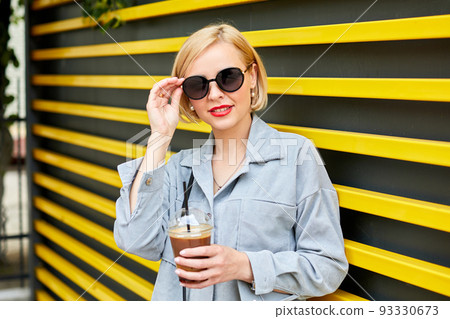 Stylish young blonde in sunglasses walks around the city and drinks a cold drink Stylish young blonde in sunglasses walks around the city and drinks a cold drink 93330673