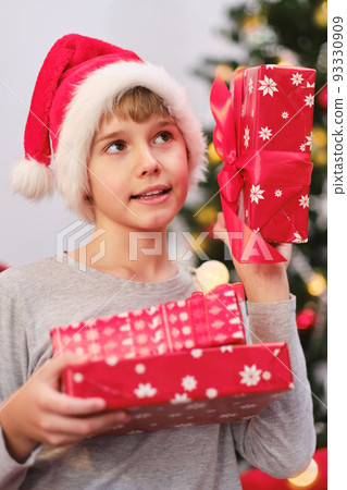 Portrait of a curious child holding presents by the Christmas tree on Christmas morning. 93330909