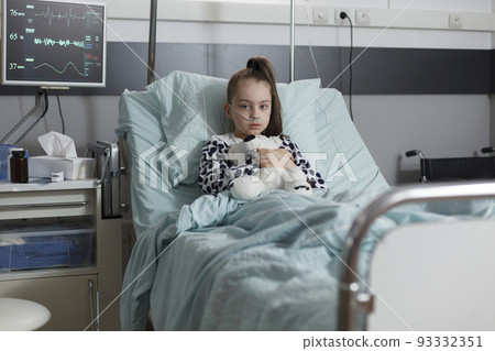Ill kid resting in children healthcare facility patient bed alone while having teddy bear. Sick little girl under treatment wearing oxygen tube while holding plush bear toy. Ill kid resting in children healthcare facility patient bed alone while having teddy bear. Sick little girl under treatment wearing oxygen tube while holding plush bear toy. 93332351