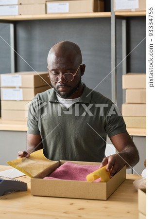 African warehouse worker packing order of customer in cardboard box while sitting at table in storage room 93334756