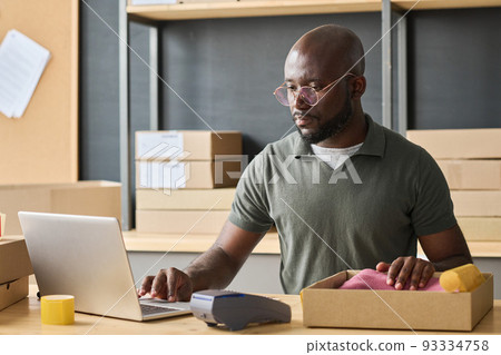 African serious man packing order in box and typing data of parcel in laptop while sitting at table in warehouse 93334758