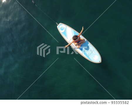 Aerial drone view on well looking middle aged woman with black hair in red swimsuit, swimming on sup in calm azure sea. Summer holiday vacation and travel concept. 93335582