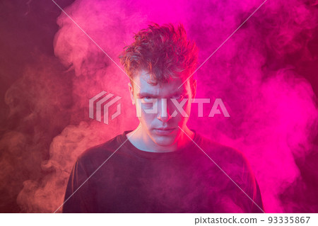 Smoke enveloped young man. Model looking at camera isolated over magenta background with clouds of smoke. Concept of mental health, art, emotions Smoke enveloped young man. Model looking at camera isolated over magenta background with clouds of smoke. Concept of mental health, art, emotions 93335867