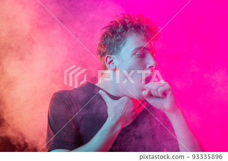 Suffocate in the smoke. Scared young man with stressed emotions isolated over pink background with clouds of smoke. Concept of facial expression, emotions Suffocate in the smoke. Scared young man with stressed emotions isolated over pink background with clouds of smoke. Concept of facial expression, emotions 93335896