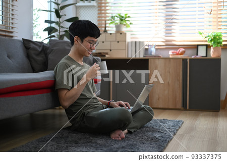 Asian man in casual clothes sitting on carpet in cozy living room and browsing internet with laptop Asian man in casual clothes sitting on carpet in cozy living room and browsing internet with laptop 93337375