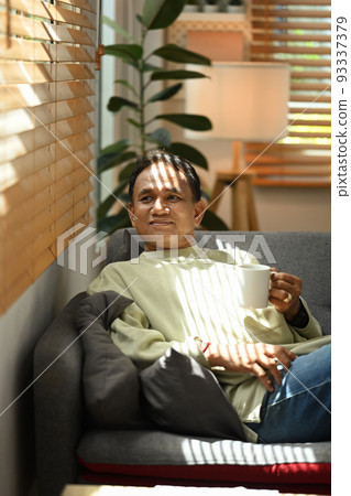 Happy senior man drinking hot coffee and relaxing on couch, enjoying stress free peaceful mood in an autumn day Happy senior man drinking hot coffee and relaxing on couch, enjoying stress free peaceful mood in an autumn day 93337379