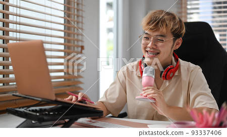 Smiling asian male using microphone and laptop computer streaming from his home studio Smiling asian male using microphone and laptop computer streaming from his home studio 93337406