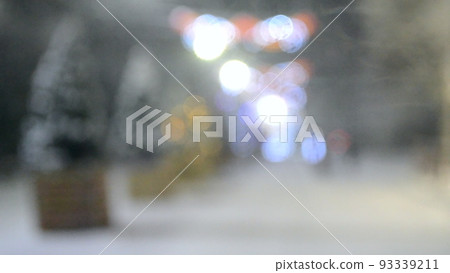 Blurred background of snowy city street with Christmas trees and illumination decoration during snowfall on winter night. People walking down street. Abstract Christmas New Year and winter background. 93339211