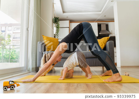 Side view of little baby boy repeating for his mother practicing yoga on yellow mat at home 93339975