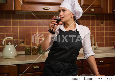 Beautiful dark-haired multi-ethnic confident woman, pleasant housewife wearing white chef's cap and black apron drinking red wine, enjoying cooking at home on her day-off. Domestic life, relaxation Beautiful dark-haired multi-ethnic confident woman, pleasant housewife wearing white chef's cap and black apron drinking red wine, enjoying cooking at home on her day-off. Domestic life, relaxation 93340548