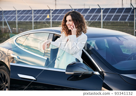 Successful business woman using 4g wireless in roaming for making international conversation and discussing solar panels, cheerful female owner of power station calling via cellphone smiling near car. 93341132