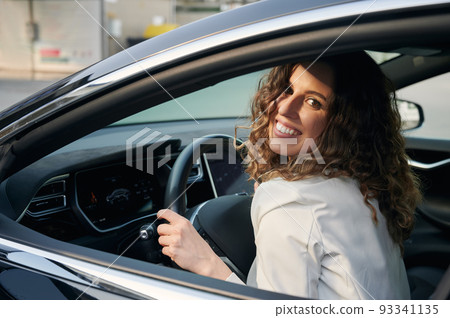 Portrait of business woman with sincere smile on face who loving her car. Curly joyful girl sitting in cab of her auto, holding hands behind the wheel. Portrait of business woman with sincere smile on face who loving her car. Curly joyful girl sitting in cab of her auto, holding hands behind the wheel. 93341135