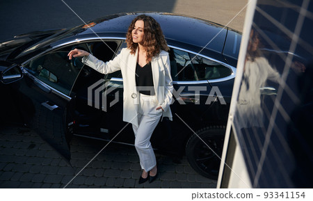 Happy woman standing by electric car on the street. Cheerful young lady with closed eyes wearing stylish white suit while posing near electric automobile outdoors. 93341154