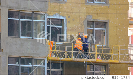 Workers are insulating the wall of a building. Construction site with working men on the elevator. panels. Hanging suspended cradle elevator. Workers are insulating the wall of a building. Construction site with working men on the elevator. panels. Hanging suspended cradle elevator. 93341784