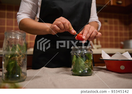 Close-up . Cropped view of a housewife in black chef's apron canning raw organic homegrown cherry tomatoes, according to traditional family recipe, in home kitchen. Marinating. Preserving. Pickling Close-up . Cropped view of a housewife in black chef's apron canning raw organic homegrown cherry tomatoes, according to traditional family recipe, in home kitchen. Marinating. Preserving. Pickling 93345142