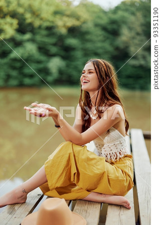 A hippie woman sits with her hat on a bridge by a lake with her hands up in the air on a nature trip and smiling happily in eco-clothing. Relaxed lifestyle 93351190