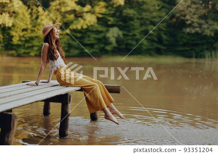 A young happy hippie woman sitting on the lake shore on a bridge in eco clothing made of natural materials in harmony with nature in the fall A young happy hippie woman sitting on the lake shore on a bridge in eco clothing made of natural materials in harmony with nature in the fall 93351204