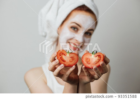 Woman holding two halves of tomato near her face with white clay mask on face 93351588