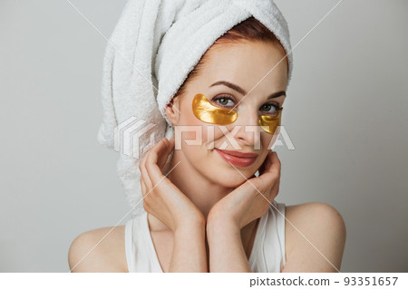 Portrait of young woman in towel touching with her hands golden hydrogel patches under eyes. Portrait of young woman in towel touching with her hands golden hydrogel patches under eyes. 93351657