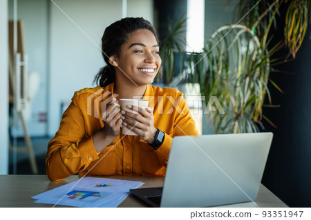 Dreamy black businesswoman using laptop and holding coffee cup, looking aside and thinking, sitting at workplace 93351947