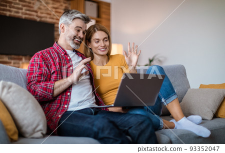 Smiling mature caucasian couple waving hand in laptop, have meeting together, sitting on sofa Smiling mature caucasian couple waving hand in laptop, have meeting together, sitting on sofa 93352047