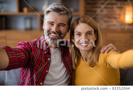 Cheerful middle aged caucasian male and female look at camera, have fun, make photo in living room interior 93352094