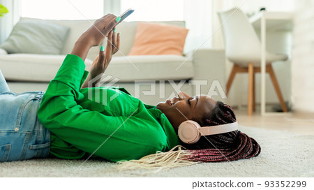 African american lady wearing wireless headphones and using smartphone, lying on floor at home, side view 93352299