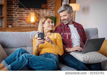 Glad adult caucasian female show phone to man with laptop, watch video in living room interior 93352761