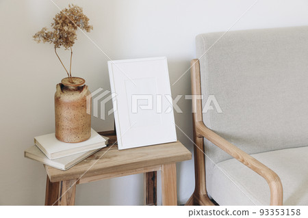 Empty vertical picture frame mockup. Ceramic vase with dry hydrangea flowers on pile of old books. Midcentury linen sofa. White wall background. Scandinavian interior, living room design. 93353158