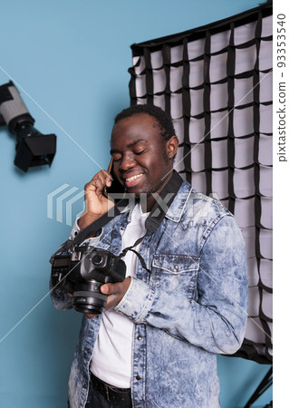 Confident photography enthusiast having a phone call in professional production studio on blue background. Happy photographer talking on smartphone while having a photo shooting. Confident photography enthusiast having a phone call in professional production studio on blue background. Happy photographer talking on smartphone while having a photo shooting. 93353540