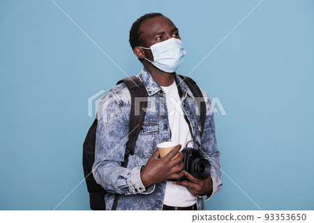 Photography enthusiast wearing virus protection facemask while having DSLR camera and backpack for holiday voyage. Young photographer wearing protective mask and having camera while going on vacation. 93353650