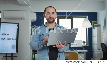 Portrait of casual business man holding laptop typing on keyboard and smiling at camera in busy modern office. Confident startup employee working relaxed in finance department of small company. Portrait of casual business man holding laptop typing on keyboard and smiling at camera in busy modern office. Confident startup employee working relaxed in finance department of small company. 93353714