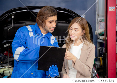 Young mechanic talking to customer asian woman about car with repairing in garage, auto service. 93359185