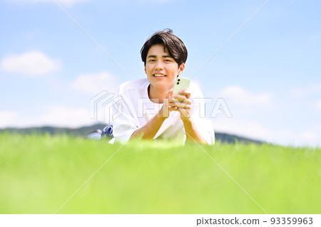 一名男子麵朝下躺在草坪上操作智能手機 一名男子麵朝下躺在草坪上操作智能手機 93359963