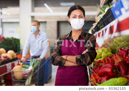 Female greengrocery owner in protective mask standing near shelves with fruits and vegetables 93362185