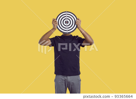 Unknown dark-skinned man hides his face behind darts board on yellow background. Unknown dark-skinned man hides his face behind darts board on yellow background. 93365664