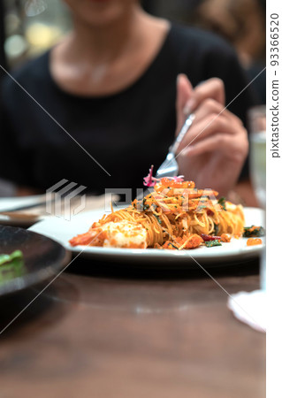 Close up and focus on the spaghetti noodle in the white dish white blur black dress female hand and her fork on it. 93366520