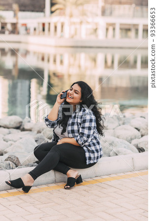 Smiling indian lady talking on phone sitting outdoors. Smiling indian lady talking on phone sitting outdoors. 93366653