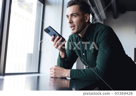 Caucasian young businessman talking on smart phone speaker by window at creative office Caucasian young businessman talking on smart phone speaker by window at creative office 93369426