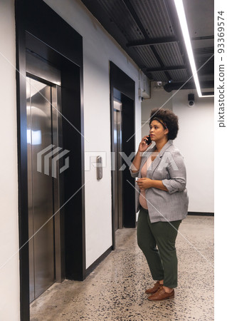 Mid adult biracial businesswoman talking on smartphone while waiting for elevator in office building 93369574
