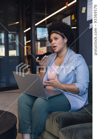 Mid adult biracial businesswoman talking on smartphone speaker while sitting with laptop in office 93369576
