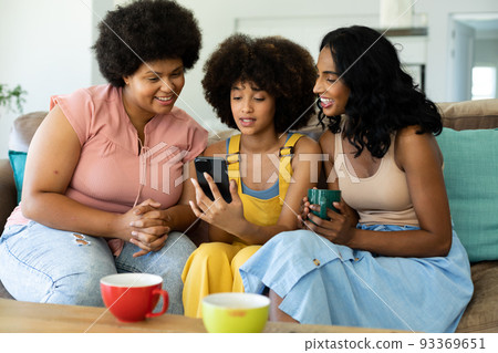 Biracial young woman using smartphone while sitting with female friends on sofa at home 93369651