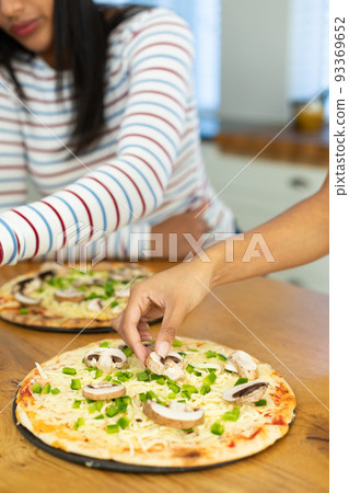 Hands of biracial female friends garnishing pizza with mushroom and bell peppers on kitchen island Hands of biracial female friends garnishing pizza with mushroom and bell peppers on kitchen island 93369652