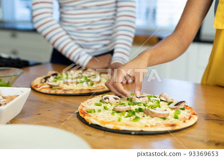 Midsection of biracial female friends garnishing pizza with mushroom and bell pepper in kitchen 93369653